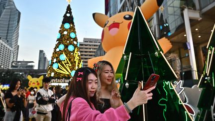 Devant un centre commercial de Bangkok, en Thaïlande, deux jeunes femmes se photographient devant des décorations de Noël à l'effigie de Pikachu, héros des Pokémon, le 25 décembre 2023. (LILLIAN SUWANRUMPHA / AFP)