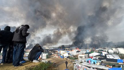 Des réfugiés regardent la fumée qui s'élève au-dessus des abris de fortune en feu dans&nbsp;la "Jungle" à Calais, le 26 octobre 2016, lors du démantèlement du camp. (PHILIPPE HUGUEN / AFP)