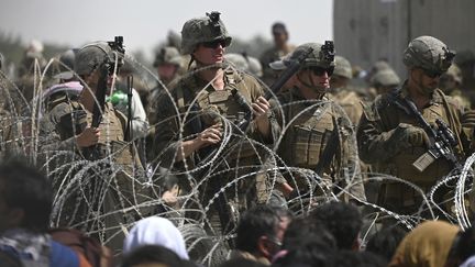 Des soldats américains montent la garde derrière des barbelés alors que des Afghans sont assis au bord d'une route à l'aéroport de Kaboul, le 20 août 2021. (WAKIL KOHSAR / AFP)