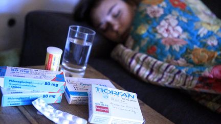 Des médicaments sur une table de nuit et une personne malade en deuxième plan. (ANGELIQUE SUREL / MAXPPP)