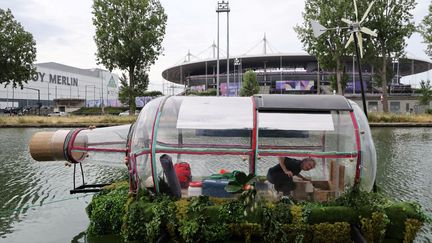 L'artiste Abraham Poincheval dans une bouteille géante fermée, sur le canal Saint-Denis, avec le Stade de France en arrière-plan, à Saint-Denis (Seine-Saint-Denis), le 25 juillet 2024. (LUDOVIC MARIN / AFP)