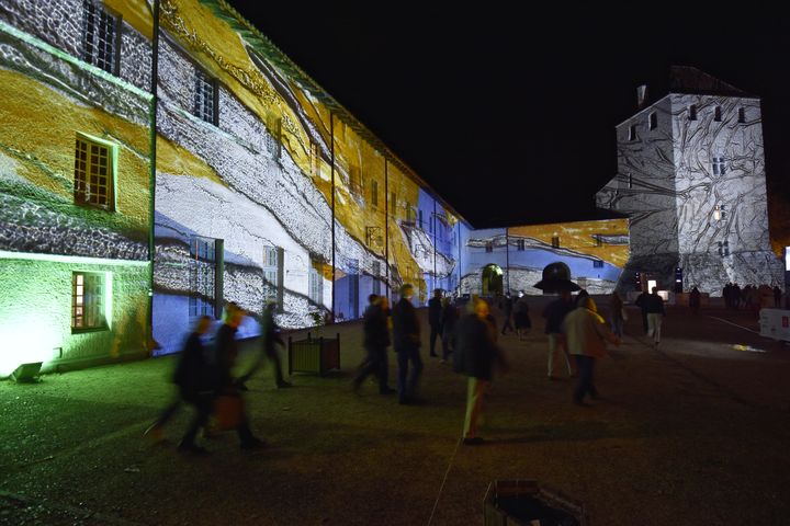 Les festivaliers traversent la cour de l'Abbaye d'Ambronay, dont les façades sont couvertes par la projection lumineuse d'une oeuvre, "Lumière de verre",&nbsp;signée par le maître verrier Julien Guiler. (BERTRAND PICHENE / FESTIVAL D'AMBRONAY)