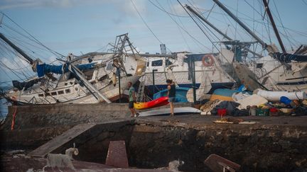 Un couple porte une embarcation, longeant des dizaines de bateaux endommagés à Dzaoudzi, à Mayotte, le 17 décembre 2024. (DIMITAR DILKOFF / AFP)