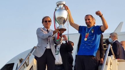 L'entraîneur italien Roberto Mancini et le défenseur Giorgio Chiellini brandissent le trophée Henri-Delaunay à leur arrivée à Rome le 12 juillet 2021 au lendemain de leur victoire en finale face à l’Angleterre. L’Italie vient de remporter son premier titre continental depuis 1968 et ce seulement trois ans après avoir manqué la Coupe du Monde 2018.&nbsp; (AEROPORTO DI ROMA / AFP)