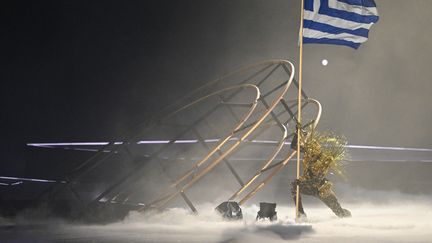 A côté d'un des anneaux enfouis, le "Golden Voyager" plante le drapeau grec qui lui a été remis par "Yamakasi" en rappel de la cérémonie d'ouverture. Il découvre ainsi l'histoire et l'esprit des Jeux olympiques. (JUNG YEON-JE / AFP)