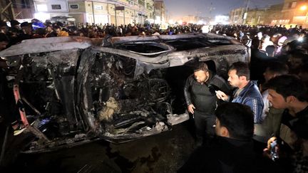 Des passants devant la carcasse d'un véhicule détruit par une frappe de drone, le 7 février 2024 à Bagdad (Irak). (MURTADHA AL-SUDANI / ANADOLU / AFP)