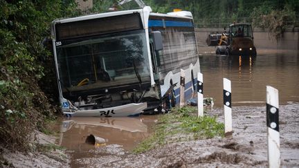 Un bus gît en travers d'une route totalement submergée par l'eau et la boue à Euskirchen, en Allemagne, le 16 juillet 2021. Derrière lui, un engin de chantier tente de dégager la route.&nbsp; (SEBASTIEN BOZON / AFP)