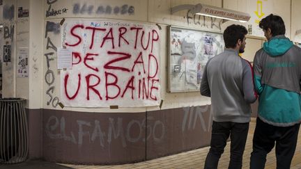 Deux hommes, dont un livreur Deliveroo, discutent dans un couloir du site de Tolbiac devant une affiche "Start-up de zad urbaine" le 13 avril 2018.&nbsp; (ARTHUR HERVE/REA)