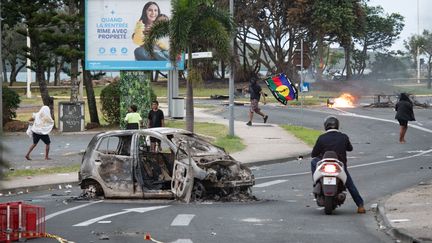 Des personnes marchent autour d'une carcasse de voiture incendiée, le 14 mai 2024 à Nouméa, en Nouvelle-Calédonie. (DELPHINE MAYEUR / AFP)