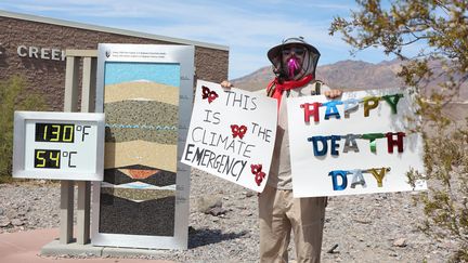 Un activiste tient deux pancartes "C'est l'urgence climatique" et "Bonne journée de la mort" devant un panneau indiquant une température de 54°C, dans la vallée de la Mort, en Californie, le 16 juillet 2023. (RONDA CHURCHILL / AFP)