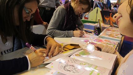 Fabienne Blanchut et Camille Dubois "Princesse parfaite"v (Fleurus). Une assiette de Petits Lu et le sourire : Fabienne Blanchut et Camille Dubois échangent gaiement avec les petites filles venues chercher leur dédicace. "On aime bien faire ça toutes les deux . C’est rare de pouvoir le faire, c’est pour ça qu’on aime bien les dédicaces au salon du livre", explique Catherine Blanchut. "Ca va de la toute petite princesse qui passe sous la table, à la grande soeur qui veut compléter sa vcollection, à la mamie... C'est très agréable d'échanger. Certains parents nous font même des suggestions pour les prochains numéros !"
 (Laurence Houot/Culturebox)