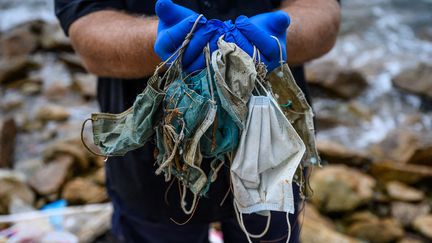 Un homme pose avec des masques jetés qu'il a trouvés sur une plage du quartier résidentiel de Discovery Bay sur l'île périphérique de Lantau à Hong Kong, le 13 mai 2020. (ANTHONY WALLACE / AFP)