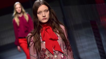 Défilé le plus attendu de la semaine de la mode milanaise, Gucci  n'a pas déçu avec une collection signée par le nouveau directeur créatif Alessandro Michele en rupture avec les saisons passées. Exit la femme de pouvoir sexy et flamboyante, place à une jeune fille romantique et androgyne. A l'entrée du théâtre de l'hôtel Diana, où Gucci  prend toujours ses quartiers, l'atmosphère était électrique. Dans un bruit tonitruant de train qui s'éloigne, le premier mannequin est apparu longeant le mur carrelé d'une station de métro au couleurs rouge sombre. Elle portait un chemisier transparent et une jupe mi-longue plissée en cuir d'un rouge vif. Seul signe distinctif: la ceinture noire barrée des deux G dorés et croisés du logo. Avec son béret ou bonnet de laine à pompon au choix, ses lunettes intello,ses robes-combinaisons à fleurs et ses chaussures plates (pantoufles fourrées trompe-l'oeil en forme de faux mocassins, sandales de moine en cuir ou fourrée et ballerines à longs lacets croisés sur les chevilles), la nouvelle femme Gucci semble avoir rajeuni de 20 ans. Dépouillée de tout accessoire clinquant, cette jeune fille dégage une certaine innocence. Non sans ironie et malice, toutefois, lorsqu'elle enfile un débardeur en dentelles à fines bretelles ou une mini robe-nuisette noire transparente. Après le départ de Frida Giannini et de Patrizio di Marco, Alessandro Michele, collaborateur de longue date de Frida Giannini et responsable jusqu'alors des accessoires Gucci, a eu la délicate mission de changer rapidement l'image de la marque afin de la relancer. Après avoir signé en janvier une première collection masculine pour Gucci, déjà remarquée, le designer a poursuivi son travail de métamorphose radicale sur la femme. Au final, comme pour l'homme, l'esprit de la collection est un brin ambigu, voire androgyne. Quand elles ne mettent pas leurs délicates robes à volant fleuries, les femmes portent le costume et se confondent de manière troublante avec les mannequins hommes, qui s'intercalent d'ailleurs sur le podium.
 (GIUSEPPE CACACE / AFP)