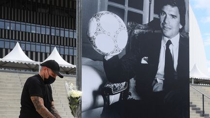 Un homme attache des fleurs sur le portrait&nbsp;de Bernard Tapie, ancien propriétaire de l'Olympique de Marseille (OM), affiché devant le stade Vélodrome à Marseille le 3 octobre 2021, après l'annonce de son décès. (CLEMENT MAHOUDEAU / AFP)