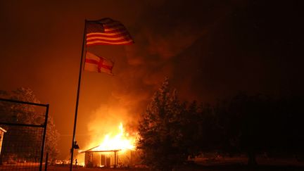 Le drapeau américain flotte devant une maison qui brûle le 23 juillet 2022 près de Mariposa en Californie. L'"Oak Fire" a détruit une dizaine de bâtiments, selon un bilan du 25 juillet 2022. (JUSTIN SULLIVAN / GETTY IMAGES NORTH AMERICA / AFP)