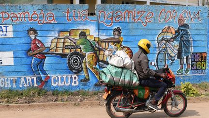 Un motard passe devant&nbsp;un&nbsp;graffiti sur un mur de&nbsp;Kibera, le plus grand bidonville de Nairobi, la capitale kényane,vsur lequel on peut lire "Pamoja tuangamize Corona", qui signifie :&nbsp;"Combattons le corona ensemble"&nbsp;(13 août 2020). (SIMON MAINA / AFP)