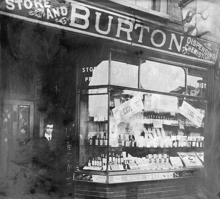 L'extérieur de la pharmacie de Burton dans les années 1900. Le commerçant se tient sur le pas de la porte. La vitrine est remplie de bouteilles, d'emballages et de publicités. (HERITAGE IMAGES / HULTON ARCHIVE/Getty Images)