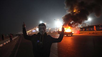 Un Egyptien portant le masque du mouvement Anonymous pose devant une voiture en flammes, &agrave; l'entr&eacute;e du stade du Caire (Egypte), le 8 f&eacute;vrier 2015. (AFP)