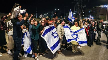 Des Israéliens brandissent des drapeaux et applaudissent après qu'un véhicule transportant des otages libérés par le Hamas soit passé vers une base militaire à Ofakim, dans le sud d'Israël, le 26 novembre 2023. (MENAHEM KAHANA / AFP)