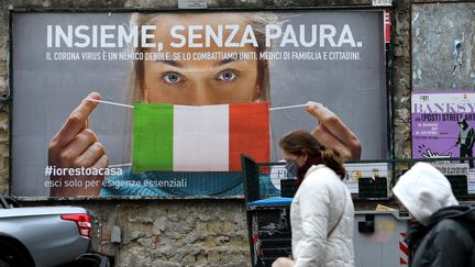 Des personnes marchent dans la rue devant une affiche incitant à se protéger avec un masque pour lutter contre le coronavirus, le 22 mars 2020, à&nbsp;Naples. (CARLO HERMANN / AFP)