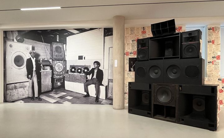 A l'exposition "Colored Black" de Dennis Morris à la Galerie du Jour, une photo en mode "wall paper" de Count Shelly,  qui importa la culture jamaïcaine du sound system à Londres, ici en 1973, à côté d'un "mur d'enceintes" de sound system. (LAURE NARLIAN / FRANCEINFO)