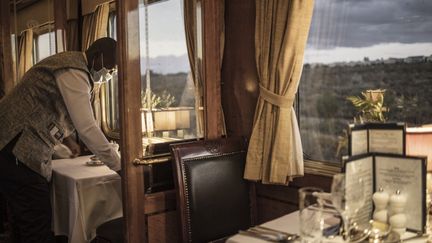 Un serveur débarrasse une table alors que le paysage semi-désertique du Karoo, en Afrique du Sud, se dévoile&nbsp;depuis le Blue Train, le 25 février 2021. (MARCO LONGARI / AFP)