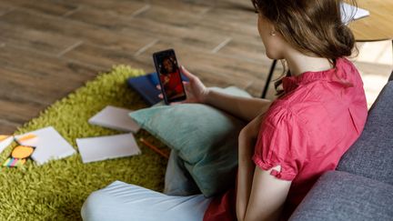 Une adolescente regarde son smartphone, assise contre un canapé. Image d'illustration. (FOTOSTORM / E+)