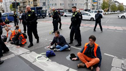 Des militants de "Dernière génération" assis sur le sol avec une main posée sur de la glue, sous le regard de policiers, dans le quartier de Schoeneberg, à Berlin (Allemagne), mardi 25 avril 2023. (ODD ANDERSEN / AFP)