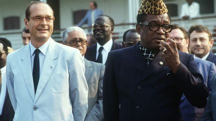 1985. Jacques Chirac est accueilli au Zaïre par le président zaïrois Mobutu Sese Seko vêtu de son «abacost». L'abacost, abréviation de «à bas le costume», est une doctrine vestimentaire imposée par Mobutu en vigueur au Zaïre entre 1972 et 1990. Afin d'affranchir la population de la culture coloniale, elle interdisait le port du costume et de la cravate, au profit d'un veston d'homme, lui-même appelé «abacost» sans col, taillé dans un tissu léger et généralement à manches courtes. Plusieurs autres pays adoptèrent ce symbole vestimentaire. (CHRISTOPHE ARCHAMBAULT / AFP Picture )