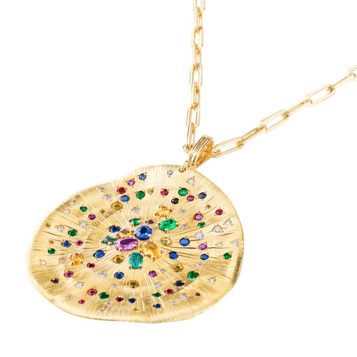 Médaille talisman soleil de Mellerio en or jaune 18k, finement gravée, cisélée et martelée. Elle est sertie de 13 saphirs jaunes, 14 saphirs bleus, 11 saphirs roses, 13 émeraudes, 14 rubis, 35 diamants. Elle se porte avec une chaîne faite main à maille rectangulaire et section trèfle (Courtesy of Mellerio)