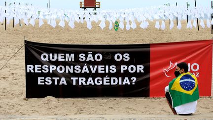 Une affiche sur laquelle est écrit "qui est responsable de cette tragédie ?", devant des tissus blancs en hommage aux Brésiliens morts du Covid-19, le 8 octobre 2021 à Rio de Janeiro (Brésil).&nbsp; (WILTON JUNIOR / ESTADÃO CONTEÚDO / AFP)