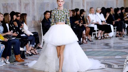 Après quelques robes cocktail courtes, en macramé, entièrement brodées de motifs floraux, le défilé Giambattista Valli fait place à des robes aux traînes majestueuses. Les jupes restent courtes à l'avant mais s'étirent à l'infini à l'arrière. Les femmes sont tantôt danseuses, portant robes de tulle et ballerines, tantôt vestales en robes drapées.
 (Patrick KOVARIK / AFP)