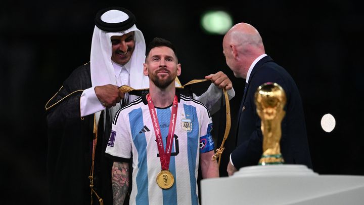 Lionel Messi, dont c'était le dernier Mondial, peut contempler le seul trophée qui manquait à son immense carrière. Avant de le brandir, l'émir du Qatar pose sur ses épaules un habit traditionnel sous les yeux du président de la Fifa Gianni Infantino. (PAUL ELLIS / AFP)