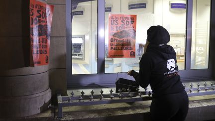 Une militante de la Fondation Abbé Pierre colle des affiches d'une campagne contre les dispositifs anti-sdf sur la façade d'une banque, le 6 décembre 2017 à Paris. (JACQUES DEMARTHON / AFP)