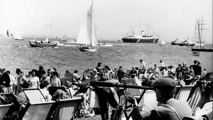 6 août 1963 à Cowes en Angleterre : Challenger potentiel pour l'America's Cup, le yacht Sovereign de 12 mètres. Les vacanciers se rassemblent sur la plage de Cowes. Le Royal Yacht Britannia peut être vu au centre. (KEYSTONE PICTURES USA / ZUMAPRESS.com VIA MAXPPP)