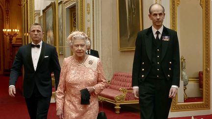 Une capture d'écran de la séquence de la cérémonie d'ouverture des Jeux olympiques de 2012 à Londres où l'on voit l'acteur britannique Daniel Craig (à gauche), jouant le rôle de James Bond, escorter la reine Elizabeth II (2e à gauche), suivie par Paul Whybrew, alias "Tall Paul", et d'un corgi, dans les couloirs du palais de Buckingham. (LOCOG / AFP)