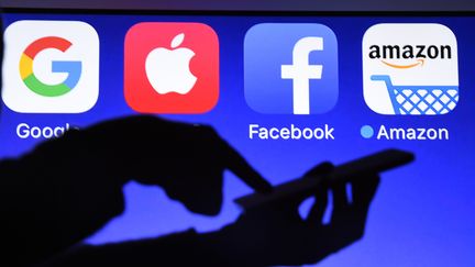 Les logos de Google, Apple, Facebook et Amazon avec à contre-jour quelqu'un qui touche un smartphone.
 (DAMIEN MEYER / AFP)