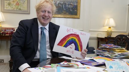 Le Premier ministre britannique Boris Johnson pose avec un dessin d'enfant lui souhaitant un prompt rétablissement, dans son bureau du 10 Downing street, à Londres, le 28 avril 2020.&nbsp; (ANDREW PARSONS / 10 DOWNING STREET / AFP)