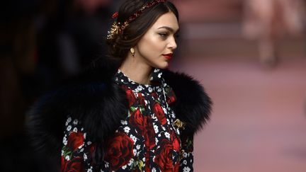 Pour Dolce &amp;Gabbana, la femme est avant tout maternelle, célébrée dans un hymne à la "Mamma". Celle que l'on vénère et que l'on veut couvrir de roses rouges, comme les fleurs à tige dorée en velours écarlate brodées, cousues, incrustées ou imprimées partout sur des robes noires en astrakan, blanches à dentelle ou encore rose tendre. "Je t'aime maman", "Tu es la plus belle maman du monde", proclament Domenico Dolce et Stefano Gabbana en lettres géantes sur robes et tricots. Dans chaque vêtement, ils déclarent un amour inconditionnel à leur mère, tout comme ils le faisaient, enfants, sur des dessins aux traits naïfs reproduits sur de gracieux ensembles. Mais attention, cette super maman, impeccable et élégante en toutes circonstances. Dans ses tailleurs années 1950, elle traverse le podium avec désinvolture, affichant ses rondeurs de femme enceinte ou tenant un sage bambin dans les bras. Certes, elle noue parfois un fichu sur la tête et utilise un cabas géant  pour faire ses courses... mais c'est un cabas en vison ou en peau de crocodile. Dans sa panoplie, aussi, un casque audio incrusté de pierreries pour écouter de la musique.
 (FILIPPO MONTEFORTE / AFP)