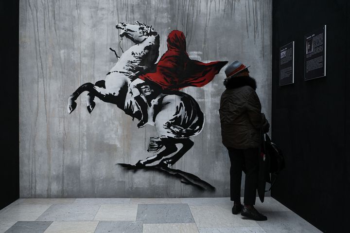 Une femme se tient près d'une reproduction d'une œuvre de Banksy (qui détourne un tableau à la gloire de Napoléon) à la gare de Milan, le 2 décembre 2021 (PIERO CRUCIATTI / ANADOLU AGENCY / AFP)