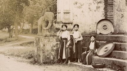 Les trois jeunes Tonkinoises portent des robes traditionnelles vietnamiennes de l'époque (áo tứ thân, littéralement «robe à quatre pans»). Deux d'entre elles se tiennent debout, adossées à la balustrade d'un temple. Derrière elles, posés çà et là, les chapeaux à large bord (nón quai thao). (Pierre-Marie Alexis Dieulefils, Hanoi, entre 1885 et 1904)