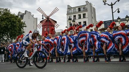 Le Français Valentin Madouas semble amusé en passant devant des danseuses du Moulin Rouge exécutant le cancan, lors de la course cycliste sur route masculine, le 3 août 2024. (MAURO PIMENTEL / AFP)