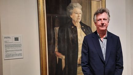 Antony Williams pose en mai 2022 devant le portrait qu'il a réalisé de la reine Elizabeth II. Il est exposé à la Mall Gallery de Londres. (RICHARD PLACE / RADIO FRANCE)