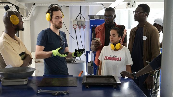 Lors d'une escale à Dakar, au Sénégal, le 22 février 2023, Baptiste Lomenech (deuxième à gauche), du projet Plastic Odyssey, présente le fonctionnement du laboratoire de transformation des déchets utilisé à bord du bateau, en vue d'encourager le déploiement de cette "low-tech" par des entrepreneurs locaux. (SEYLLOU / AFP)