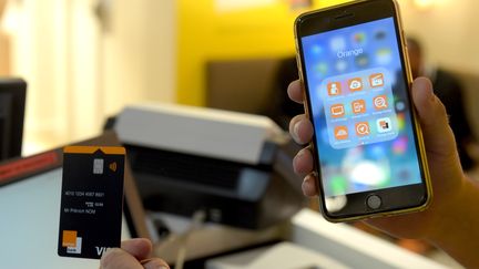 Un client tient une carte bancaire Orange Bank ainsi qu'un smartphone sur lequel est installé l'application de la banque en question, en ligne depuis le 2 novembre 2017. (ERIC PIERMONT / AFP)