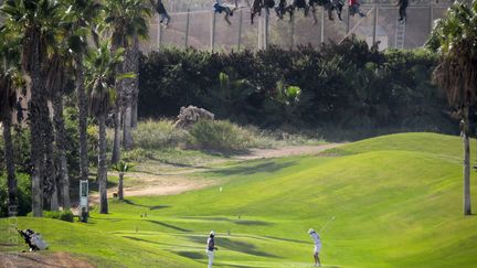 Des golfeurs progressent sur leur parcours sous le regard de migrants africains assis au sommet d'une barrière, le 22 octobre 2014 dans l'enclave espagnole de Melilla, située dans le territoire marocain. (JOSE PALAZON OSMA / PRODEIN / AFP)