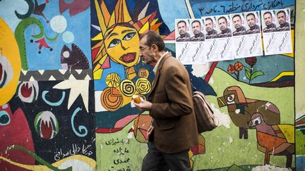 Un homme mange un beignet alors qu'il passe devant un mur peint où sont collées les affiches du candidat réformateur Kazem Jalali dans le centre-ville de Téhéran le 22 février 2016. Les leaders réformistes et modérés comptent sur leurs partisans, notamment pour faire basculer l'Assemblée des experts, qui est aujourd'hui dominée par les conservateurs. «Les ex-présidents iraniens Mohammad  Khatami (réformateur) et Akbar Hashemi Rafsandjani (modéré) ont appelé (dans des messages séparés) les électeurs à voter massivement vendredi (26 février) pour les candidats réformateurs et ainsi "barrer la route aux extrémistes"», rapporte l'AFP. Ces anciens dirigeants «sont candidats à l'Assemblée des experts dont ils souhaitent voir écartées les figures les plus conservatrices, dont son président, l'ayatollah Mohammad Yazdi». (BEHROUZ MEHRI / AFP)