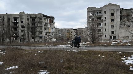 Deux personnes circulent sur une route d'Izoum le 20 février 2023, en Ukraine, devant des bâtiments partiellement détruits. (SERGEY BOBOK / AFP)