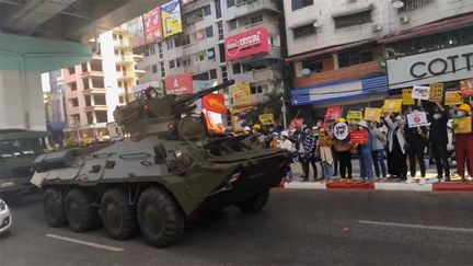 Des manifestants tenant des pancartes s'alignent sur le bord de la route alors que des chars et des camions de l'armée se dirigent vers le centre-ville de Rangoun, le 15 février 2021. (EYEPRESS NEWS / AFP)