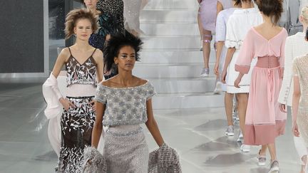 Chanel a présenté une collection fraîche, à dominante pastel dans un décor blanc. "C'est une espèce de boîte de nuit d'une autre galaxie", raconte Karl Lagerfeld. La scène tourne, apparaît un groupe de musique, mené par Sébastien Tellier, au piano. La top britannique Cara Delevingne ouvre le bal avec des tennis brodées aux pieds. Les sneakers sont tendance même en haute couture : des mannequins de Dior en portaient. La taille est près du corps, corsetée, pour mieux donner du volume à la silhouette, qui est courte et structurée. "J'ai associé ça dans ma tête à une ceinture de motocycliste", explique le couturier.. Certains looks sont accompagnés de genouillères et coudières luxe. Les corsets, avec baleines, montent haut sous la poitrine. Certains sont brodés. "Ce sont des constructions très compliquées ; on ne pourrait pas le faire en prêt-à-porter", souligne Karl Lagerfeld. Le couturier a fait travailler les ateliers appartenant à Chanel. "Il n'y a aucun tissu, à part les mousselines et les tulles qui sont faits industriellement. Et tout est fait à la main. C'est pas des trucs qu'on peut acheter. Ils font 20 centimètres par jour." "Il faut de la légèreté, sinon, la couture a une image un peu vieille. Le côté raide, y en a marre", dit-il. Au final, Cara Delevingne réapparaît, en mariée, tout en transparence, avec un garçonnet d'honneur: le filleul de Karl, Hudson, qui porte la longue traîne.
 (HENDRIK BALLHAUSEN / DPA / DPA PICTURE-ALLIANCE/AFP)